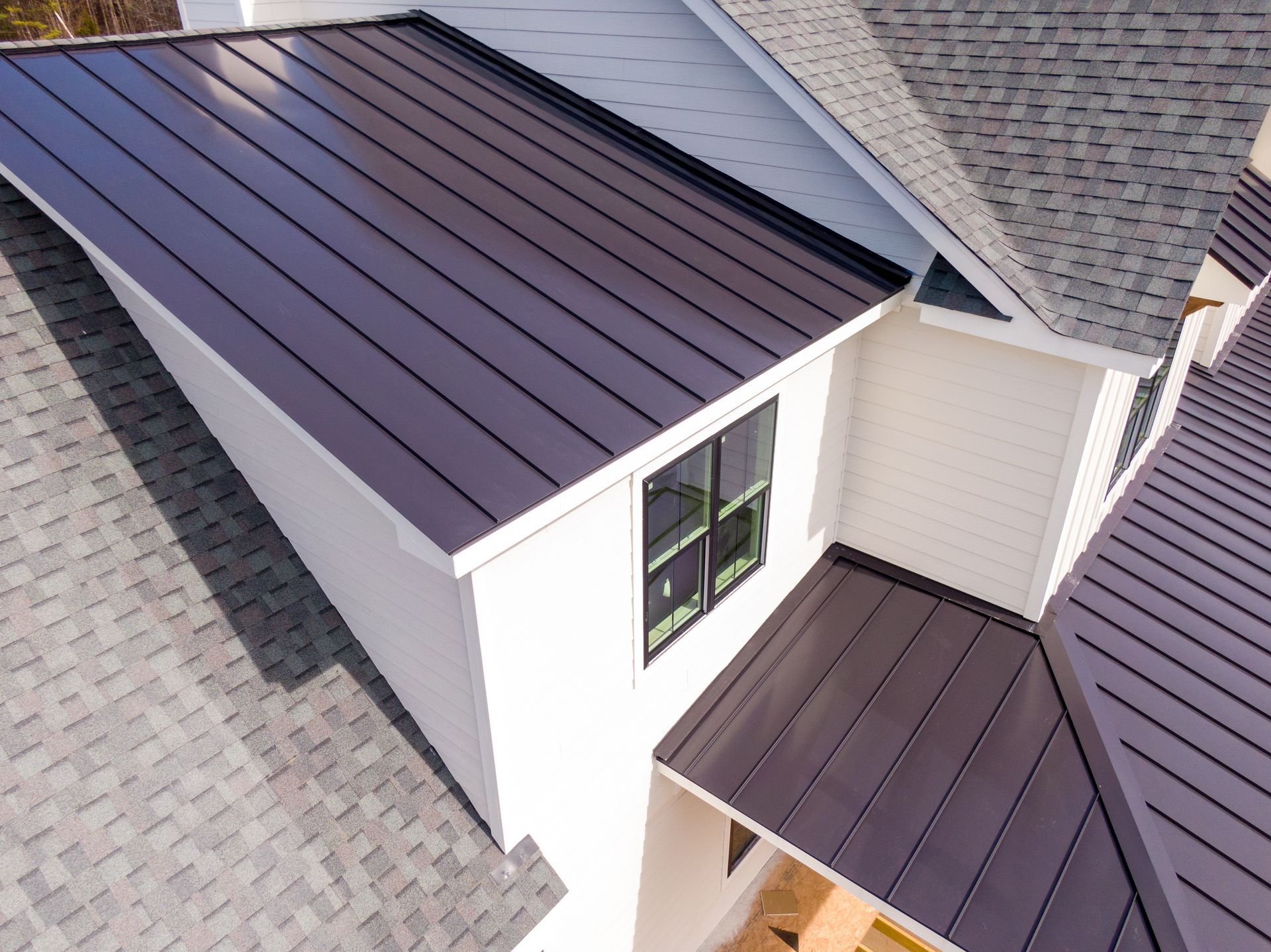 Aerial view of a house roof with dark metal panels and gray shingles around a dormer window.
