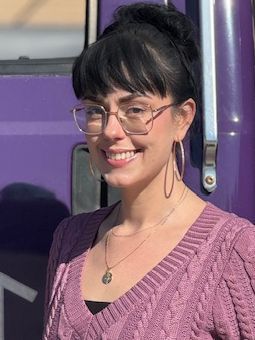 Woman with dark hair in glasses smiles in front of a purple backdrop; wearing a pink sweater and hoop earrings.