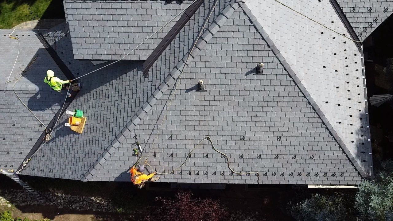 Roofers working on a gray shingled roof with several tools and safety vests visible Roofers working on a gray shingled roof with several tools and safety vests visible