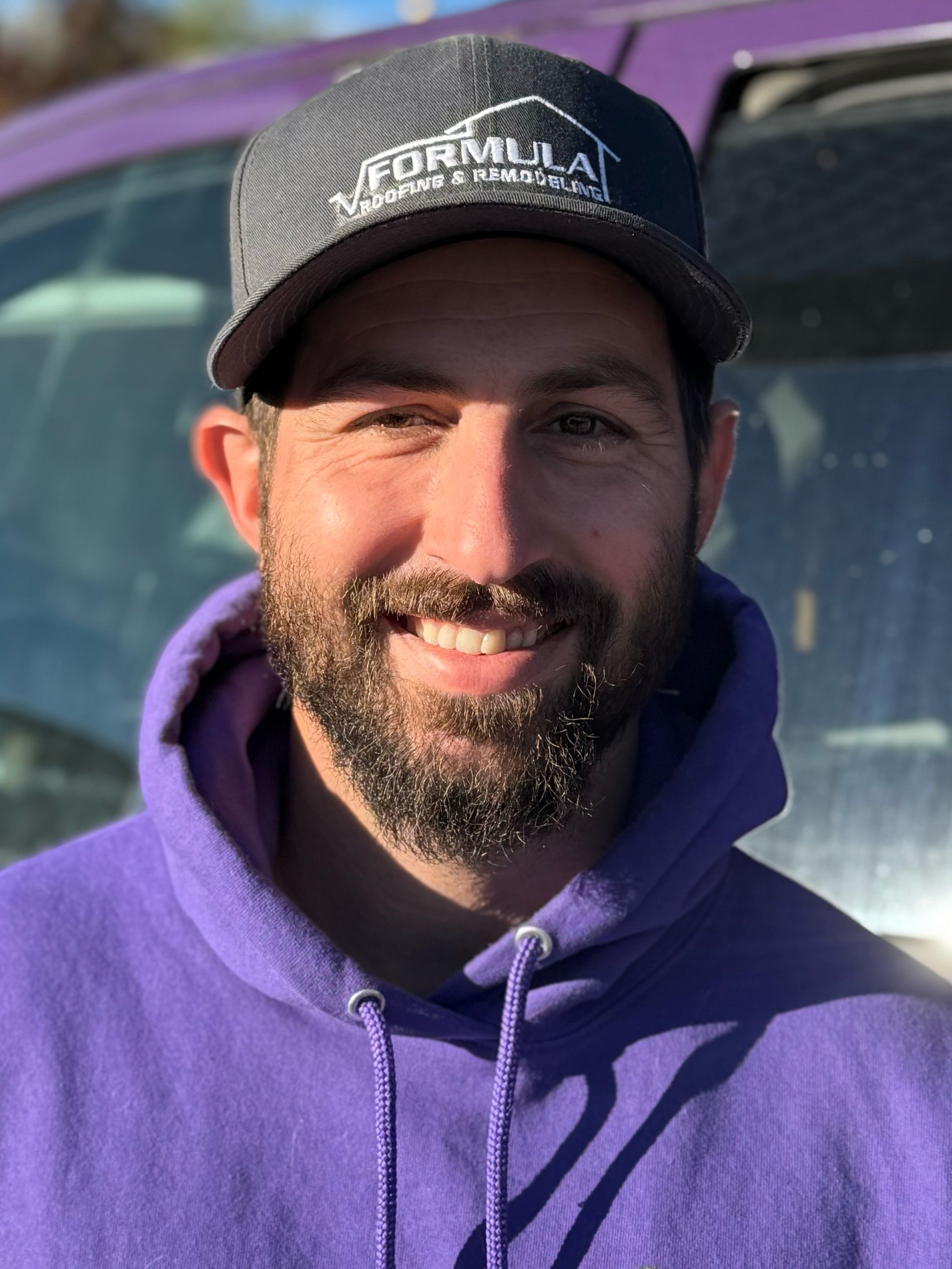 Man in a purple hoodie and cap smiles, outdoors in front of a vehicle.