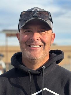 Man smiling, wearing a hat and sunglasses, a black hoodie, outdoors.