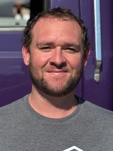 Man with a beard smiling in front of a purple vehicle. He wears a gray shirt.