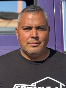 Man with short graying hair in a black shirt, looking at the camera. Purple background.