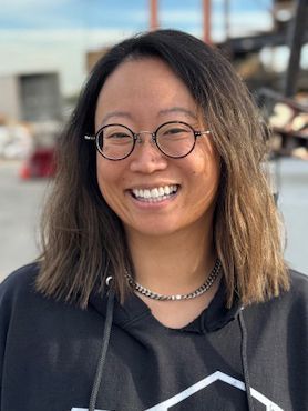 Woman with glasses and a black hoodie smiles broadly outdoors.