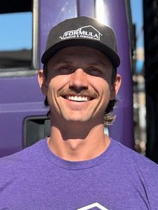 Man with mustache and Formula cap smiles in front of a purple truck.
