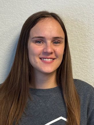 Woman with long brown hair smiles at the camera, wearing a gray sweatshirt.