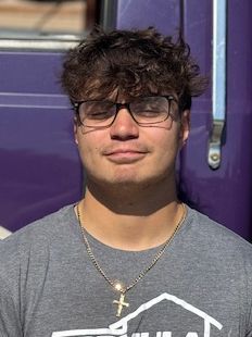 Man with glasses and curly brown hair, wearing a gray t-shirt and gold chain with a cross pendant, smiling in front of a purple backdrop.