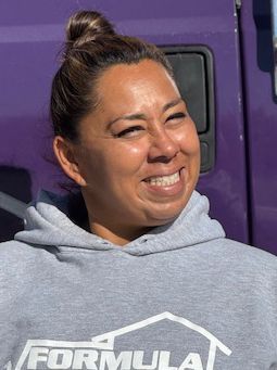 Woman in gray hoodie smiles, with a purple truck in the background. Her hair is in a bun.