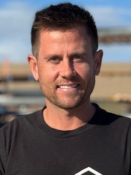 Man with short brown hair smiling, wearing a black shirt, outdoors with a building background.