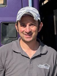 Man wearing a hat and gray shirt with logo, smiling in front of a purple vehicle.