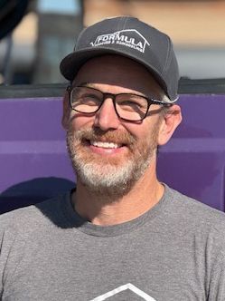 Man wearing glasses and a Formula Roofing hat smiles, against a purple background.