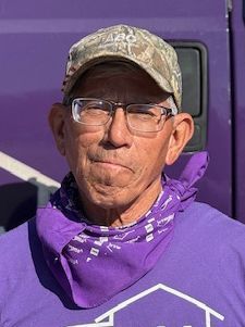 Man wearing a camo hat, glasses, and purple bandana. He is smiling and standing in front of a purple backdrop.