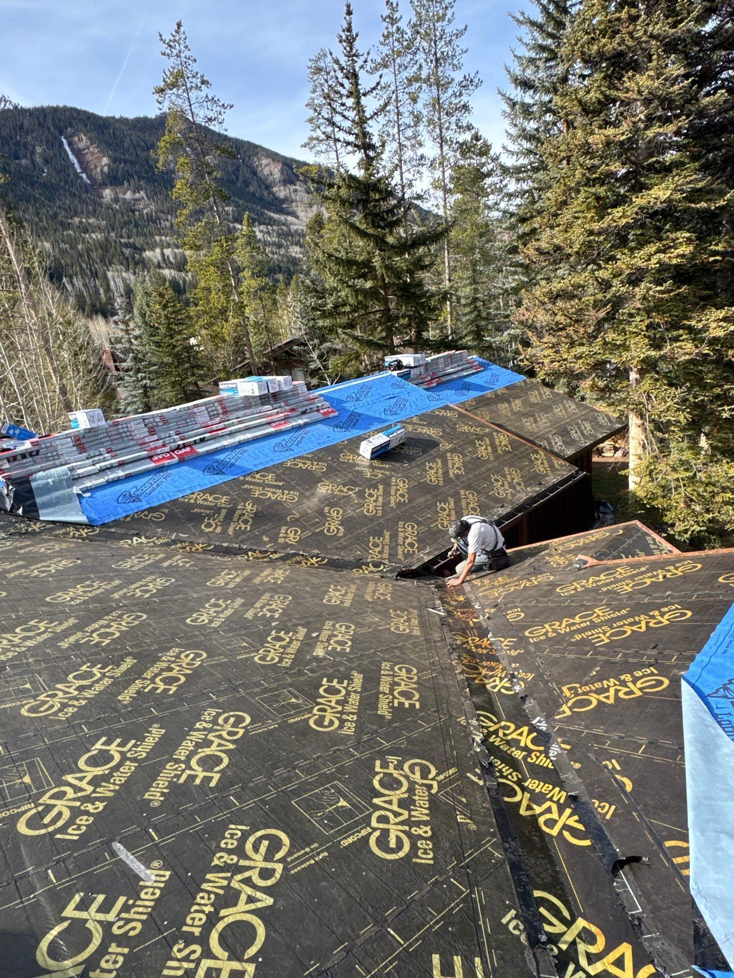 Worker laying roofing underlayment on a steep cabin roof in a forested mountain setting Worker laying roofing underlayment on a steep cabin roof in a forested mountain setting
