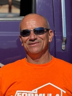 Man wearing sunglasses and an orange shirt, smiling in front of a purple vehicle.