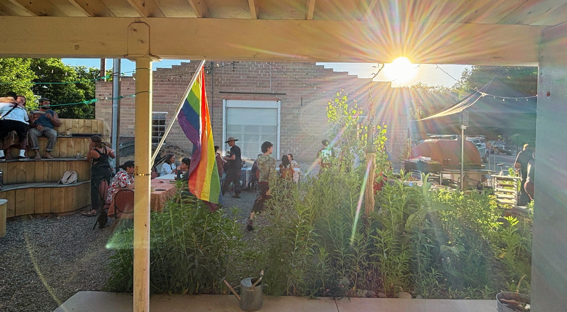 Sunlit backyard gathering with a rainbow flag, patio cover, and people near a garden