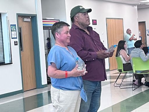 Two people standing in a cafeteria holding drinks, with tables and seated people in the background