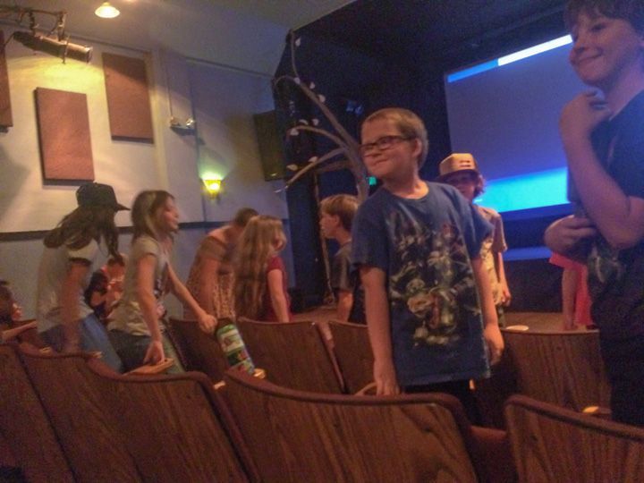Children standing in a church or auditorium, with wooden pews and a blue-lit stage in the background
