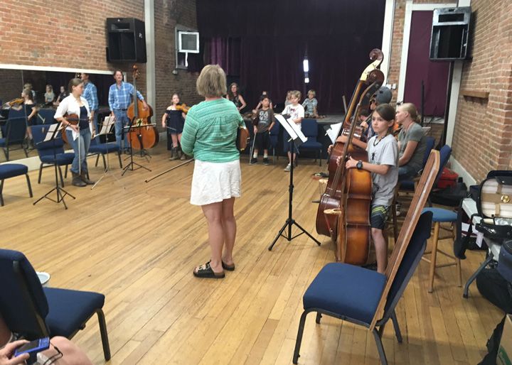 String ensemble rehearsing on a wooden-floored stage, conductor in green top and white skirt facing musicians.
