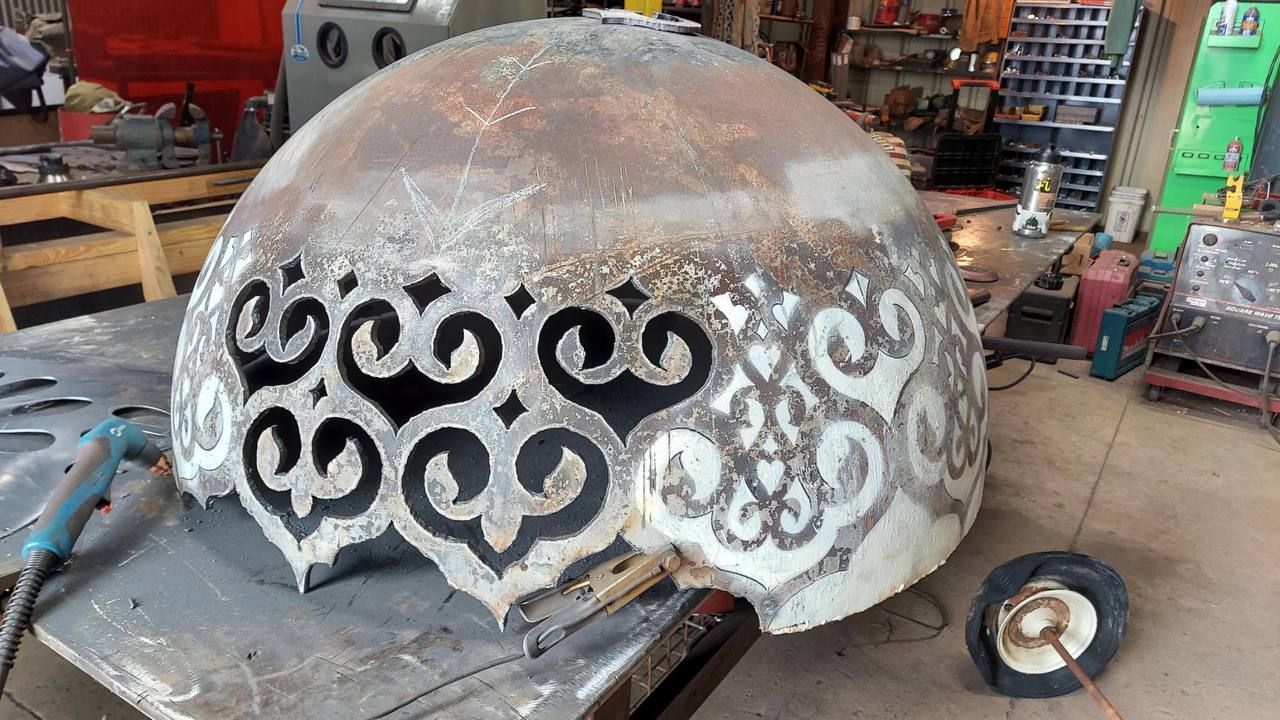 Rusty metal dome with black decorative swirl cutouts in a workshop, resting on a workbench