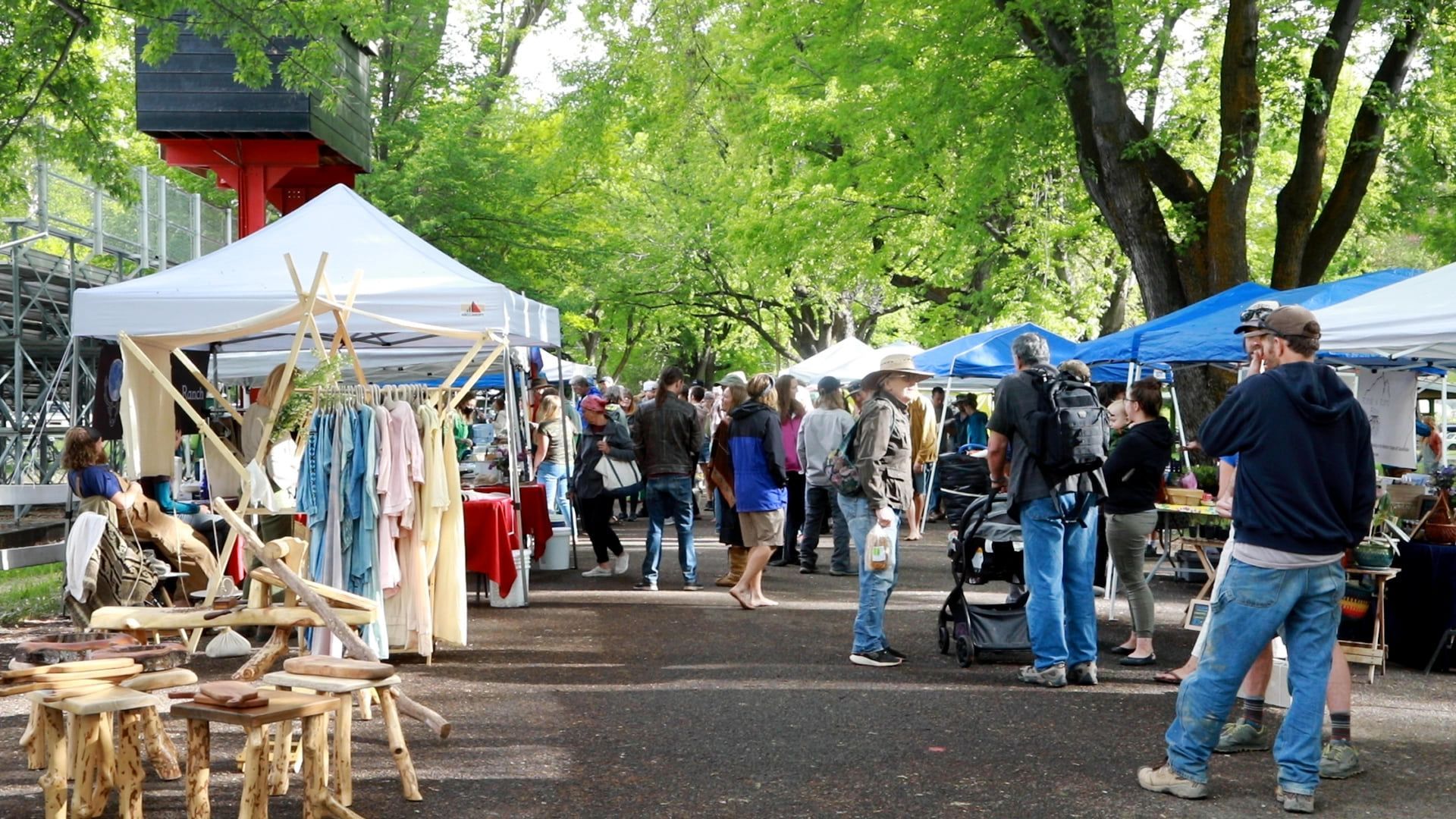 Outdoor craft market with white and blue tents, clothes racks, and people browsing along a tree-lined path