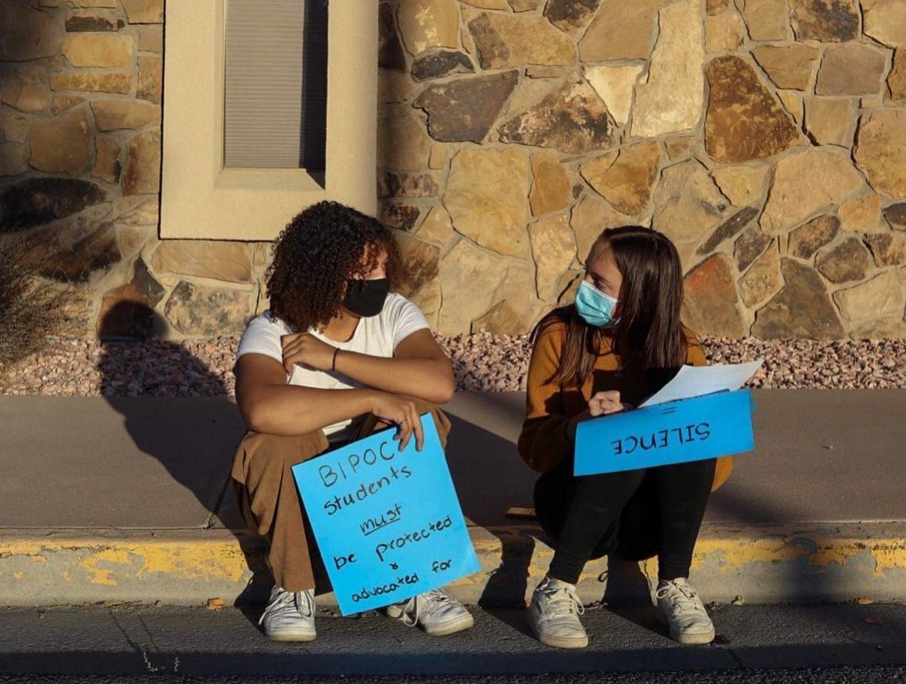 Two masked people sitting on a curb holding blue protest signs beside a stone wall.