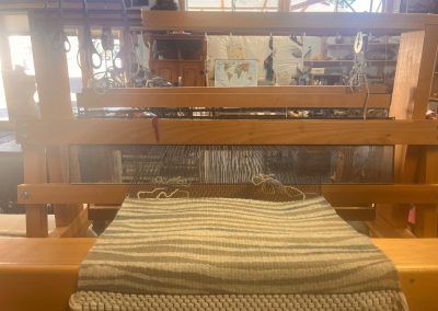 Woven textile on a wooden loom in a workshop, with threads stretched across the frame.