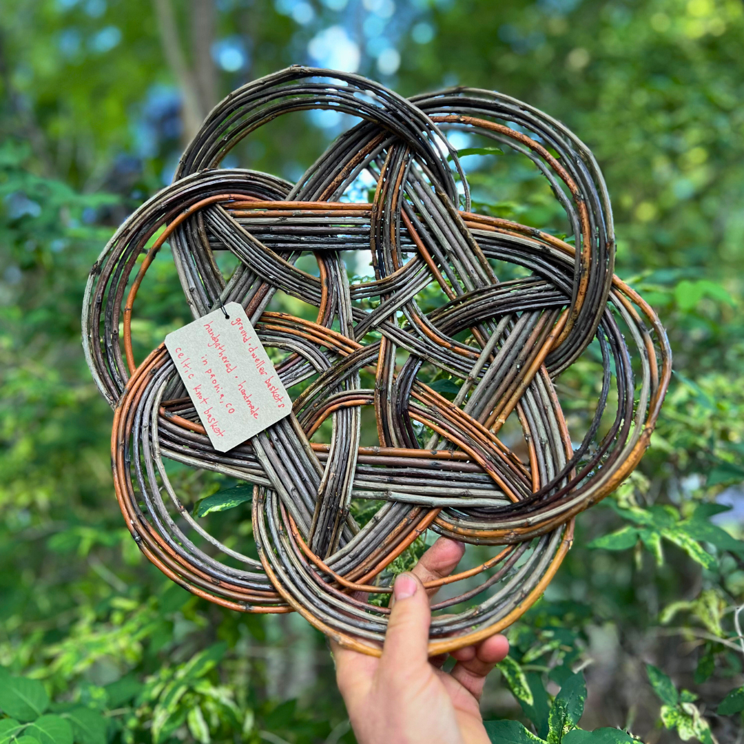 Hand holding a woven metal ring sculpture with bronze, black, and silver strands outdoors