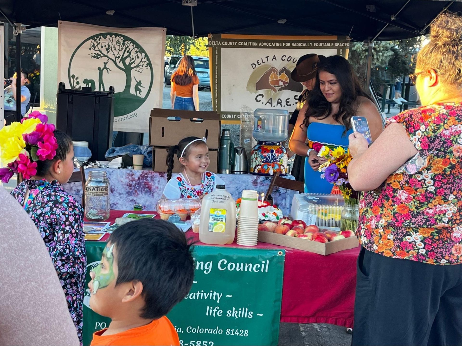 Outdoor market booth with colorful crafts, books, and a child at a table under a canopy