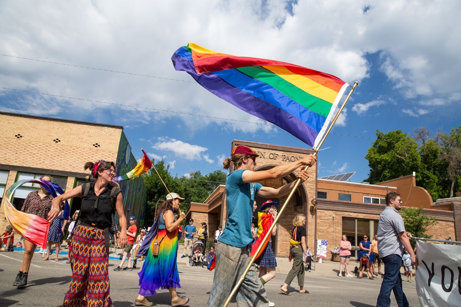 People marching with rainbow Pride flags at a street parade under a blue sky.