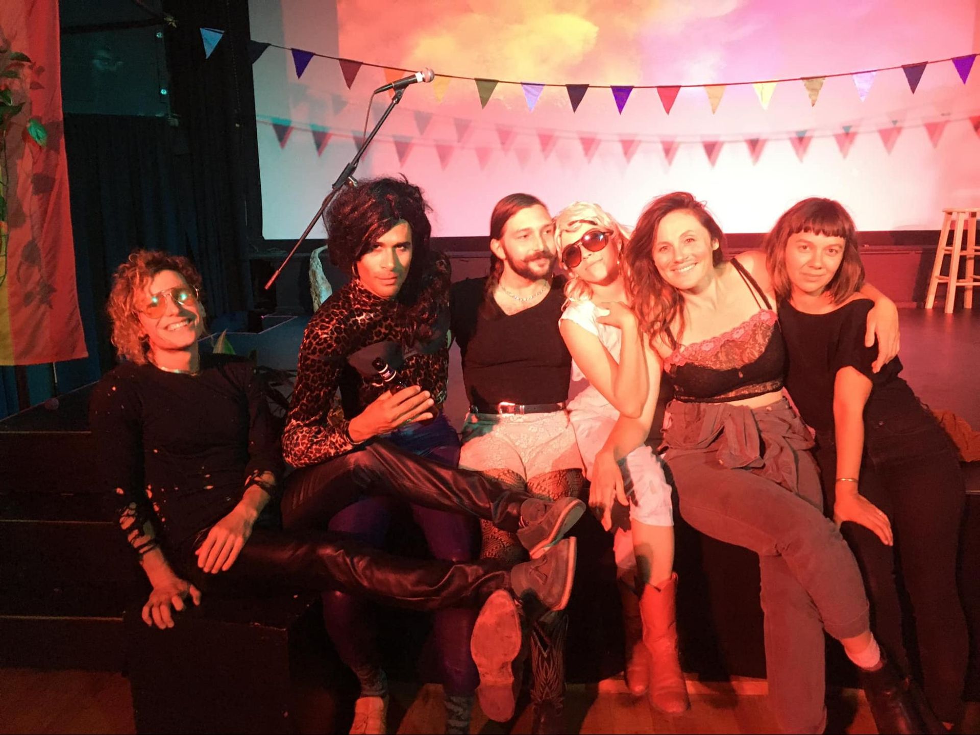 Five people sitting on a couch at a colorful party, with bunting flags and warm pink lighting.