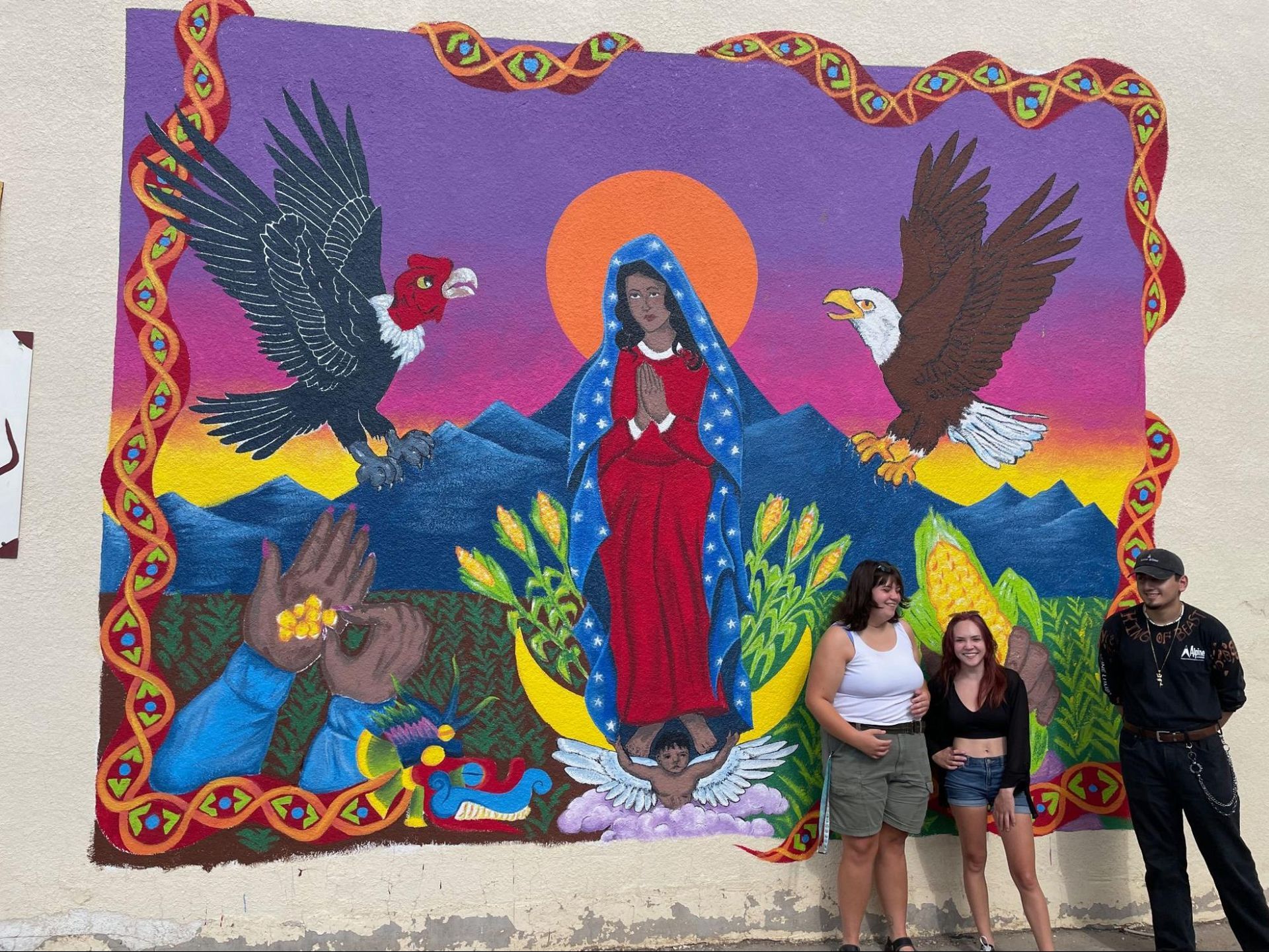 Mural of Virgin Mary with birds and mountains; three people posing in front of it.