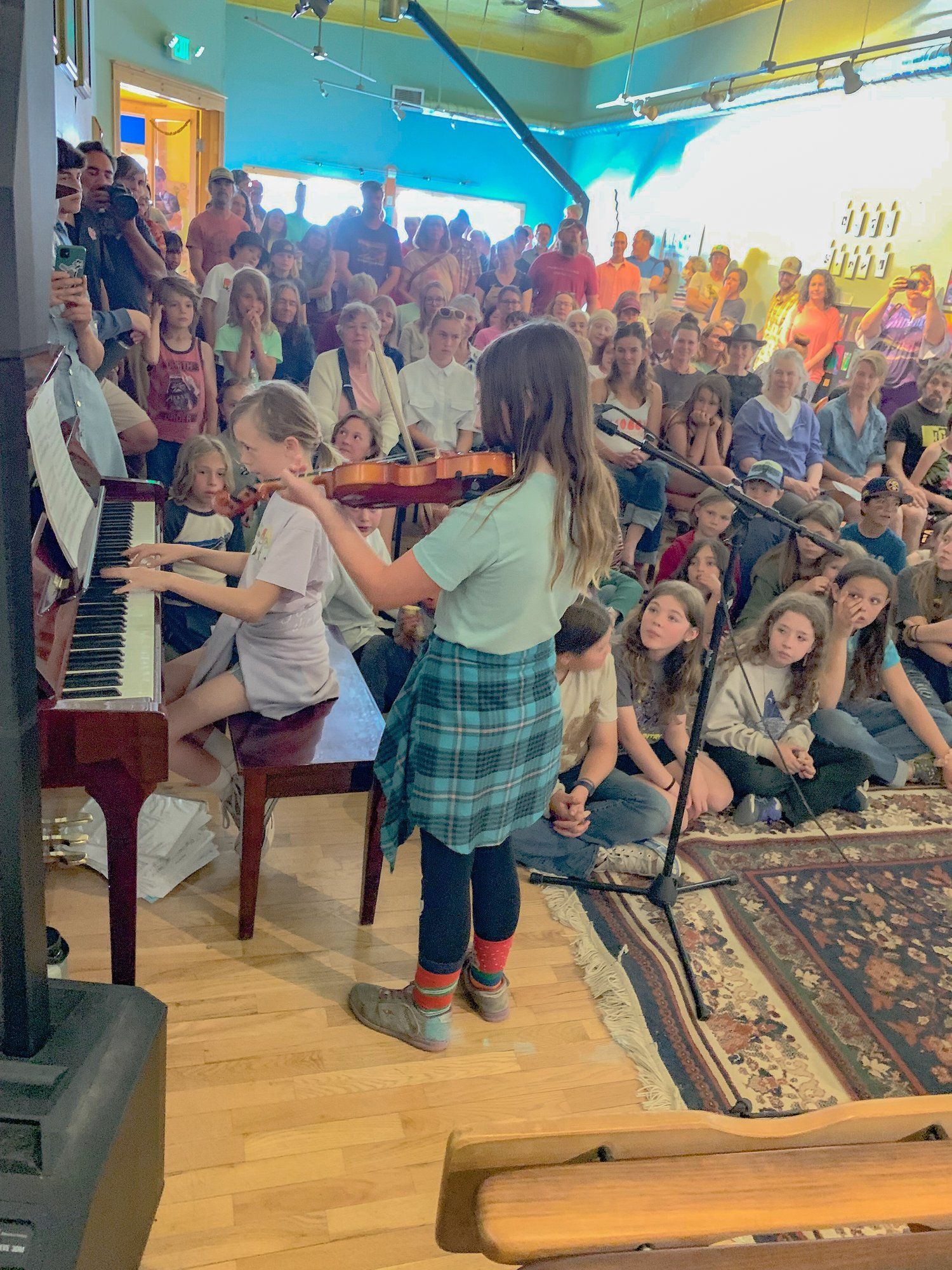 Children playing piano and guitar in a lively indoor performance with seated audience behind them