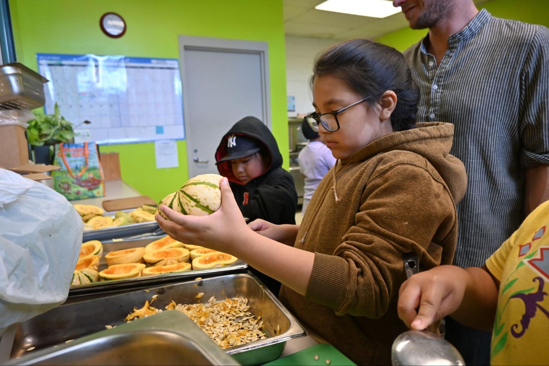 People serving food at a cafeteria counter, with a child holding a plate of fruit.