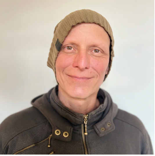 Person in a tan beanie and dark jacket, smiling softly against a plain light background