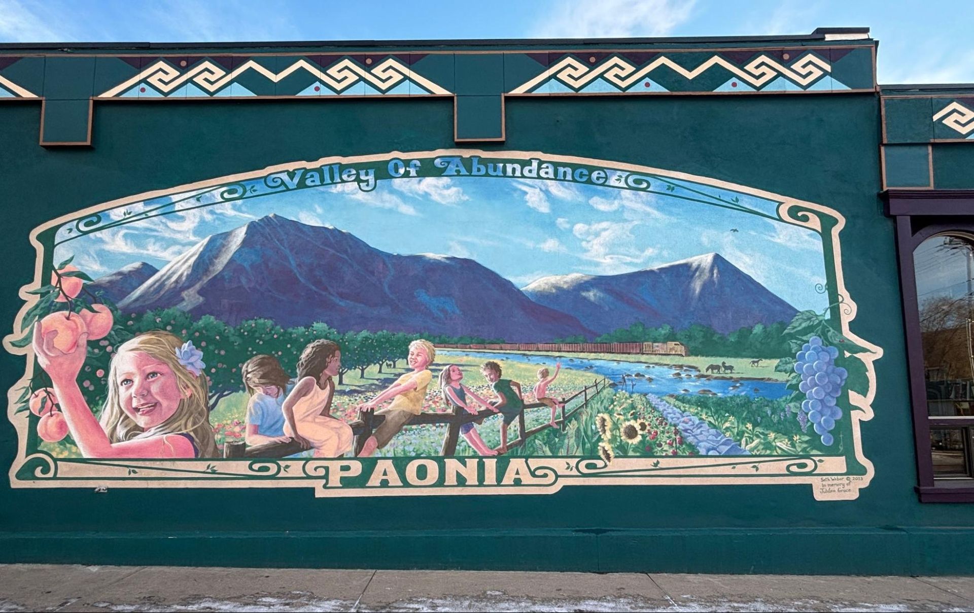 Colorful mural of a mountain valley and people on a teal wall, with the word “PAAONAO” below