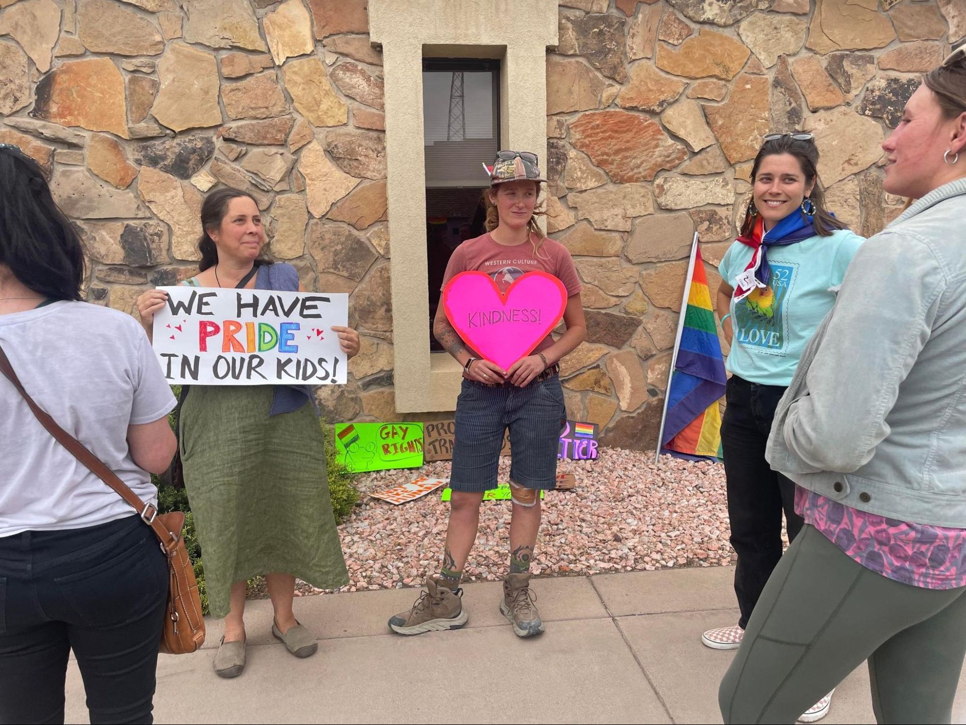 People holding signs and a pink heart, standing outside by a stone wall during a protest or rally