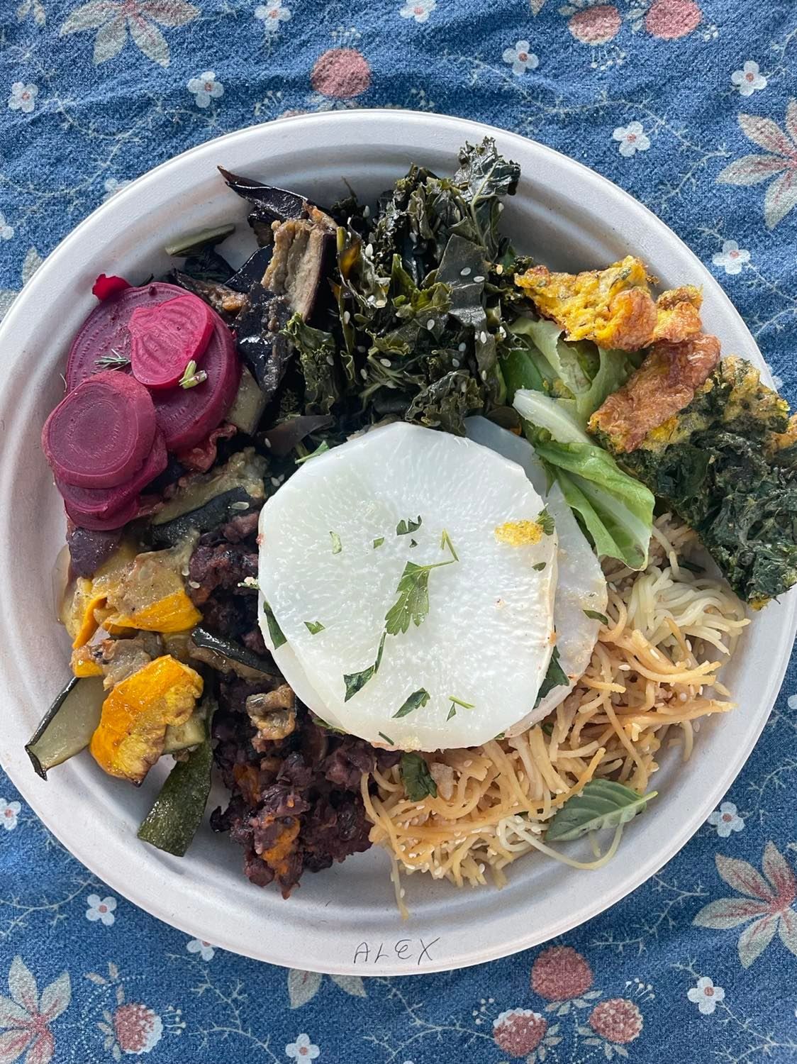 Bowl of colorful noodles and vegetables with pickled beets, greens, and a white radish slice on a blue floral tablecloth