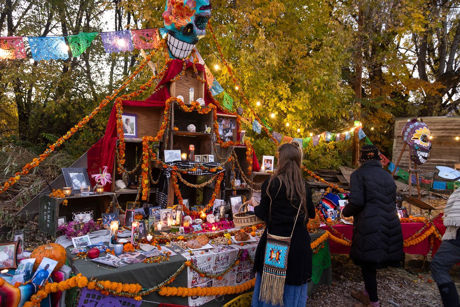 Outdoor memorial altar with candles, flowers, and offerings; two people stand nearby under autumn trees.