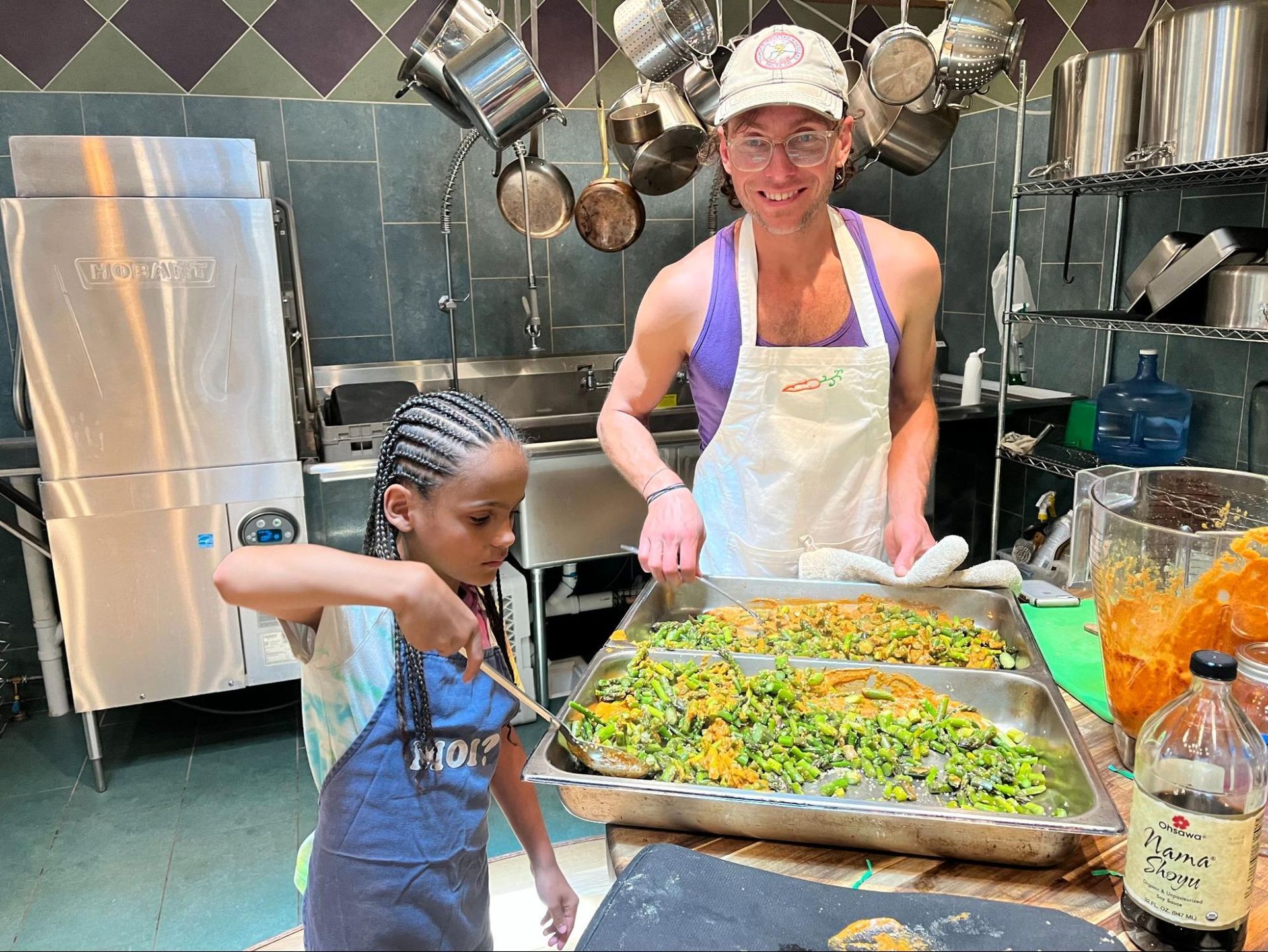 Two people in a kitchen serving a tray of roasted vegetables and fruit salad