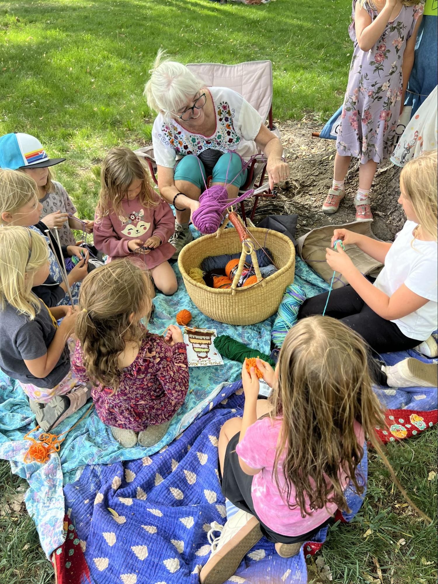 Children sit on blankets around a woven basket during an outdoor group activity in the grass.