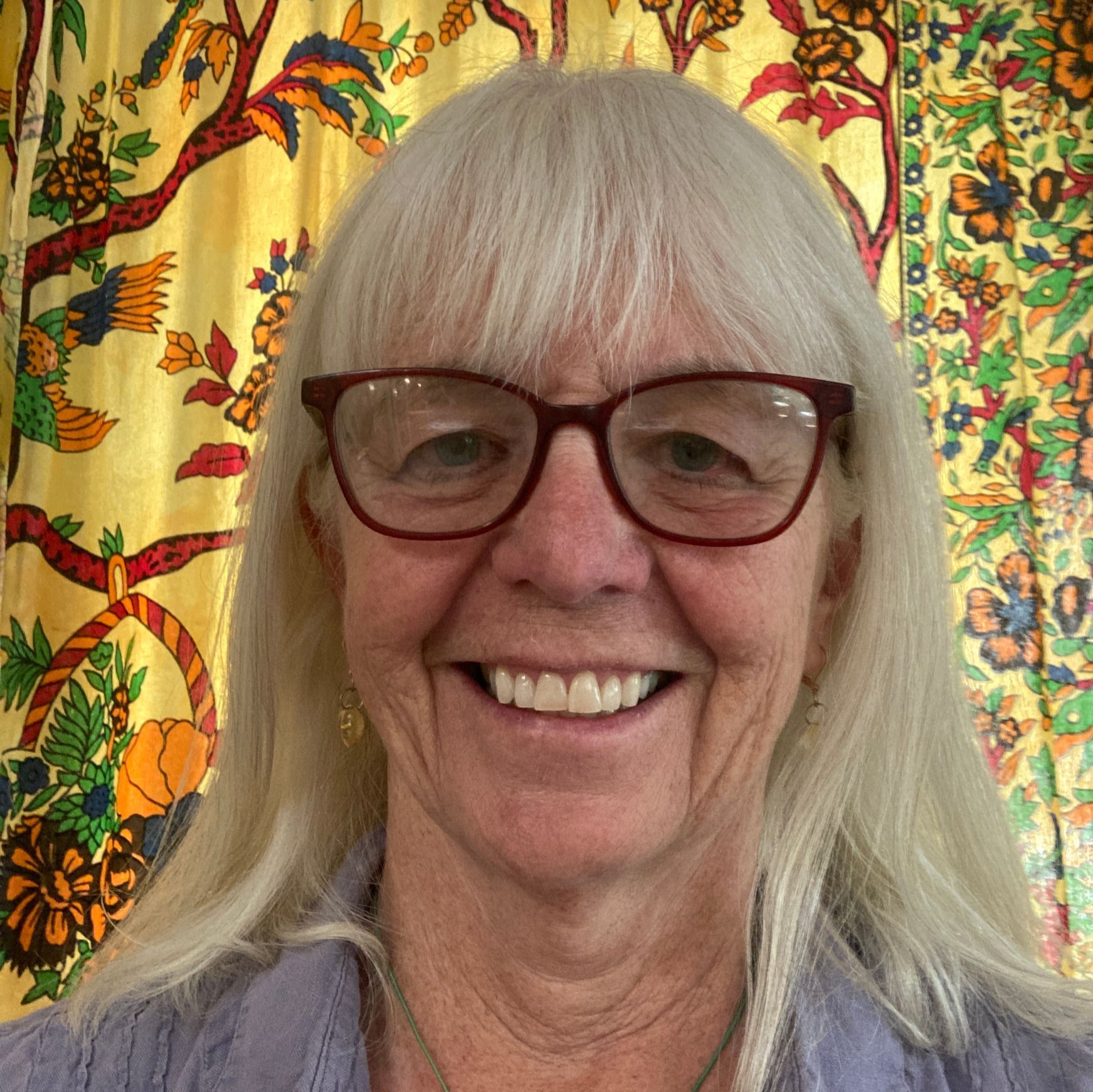 Smiling person with white hair and red glasses in front of a colorful floral tapestry