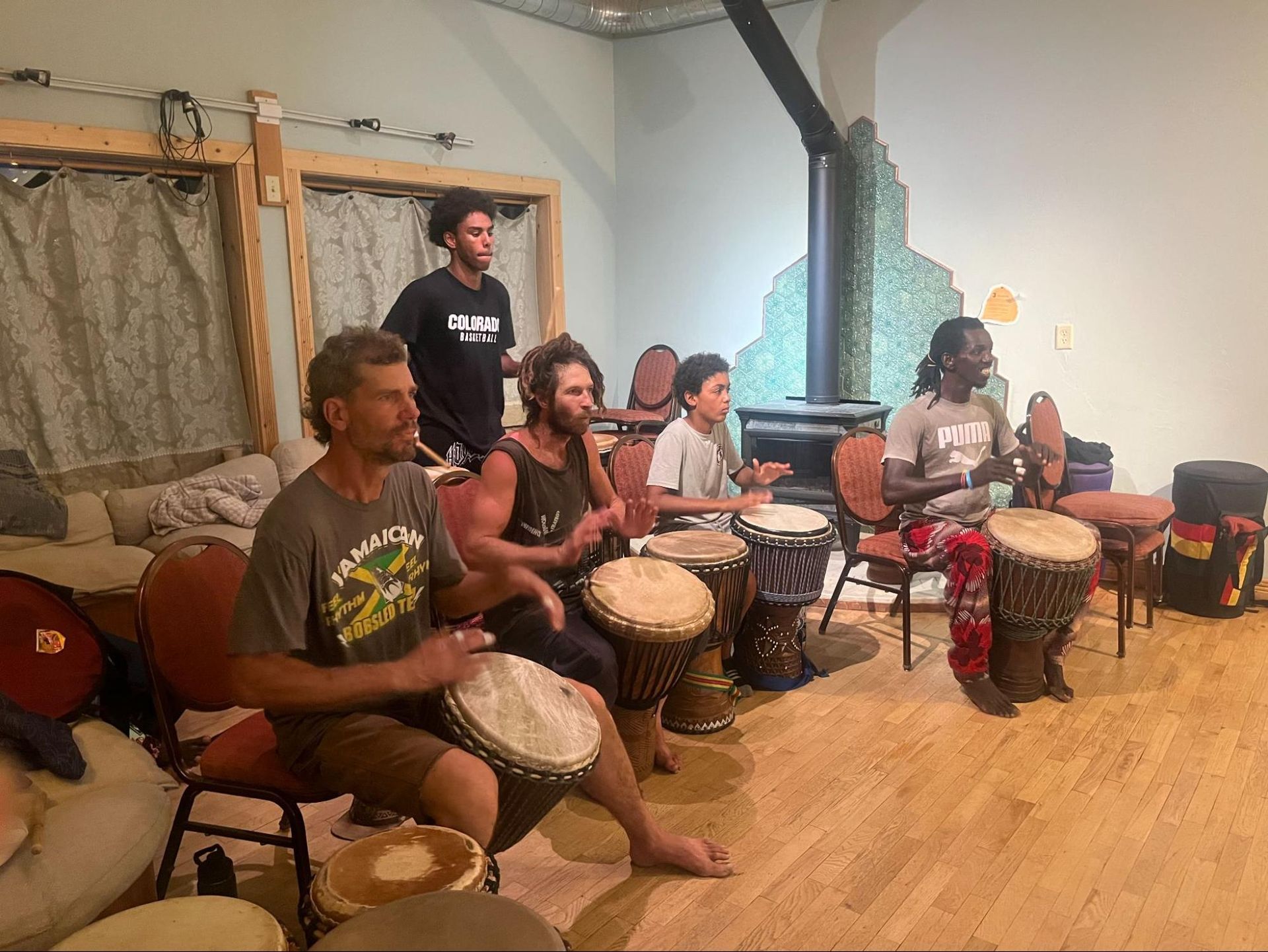 Drummers seated in a studio playing hand drums, with one person standing in the background.