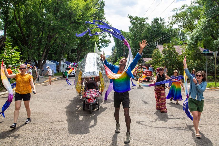 People dancing in a colorful street parade under trees, with a decorated vehicle in the center.