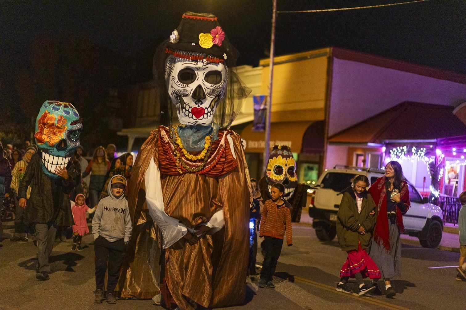 Day of the Dead parade with giant skull-costumed figures and people walking at night under streetlights