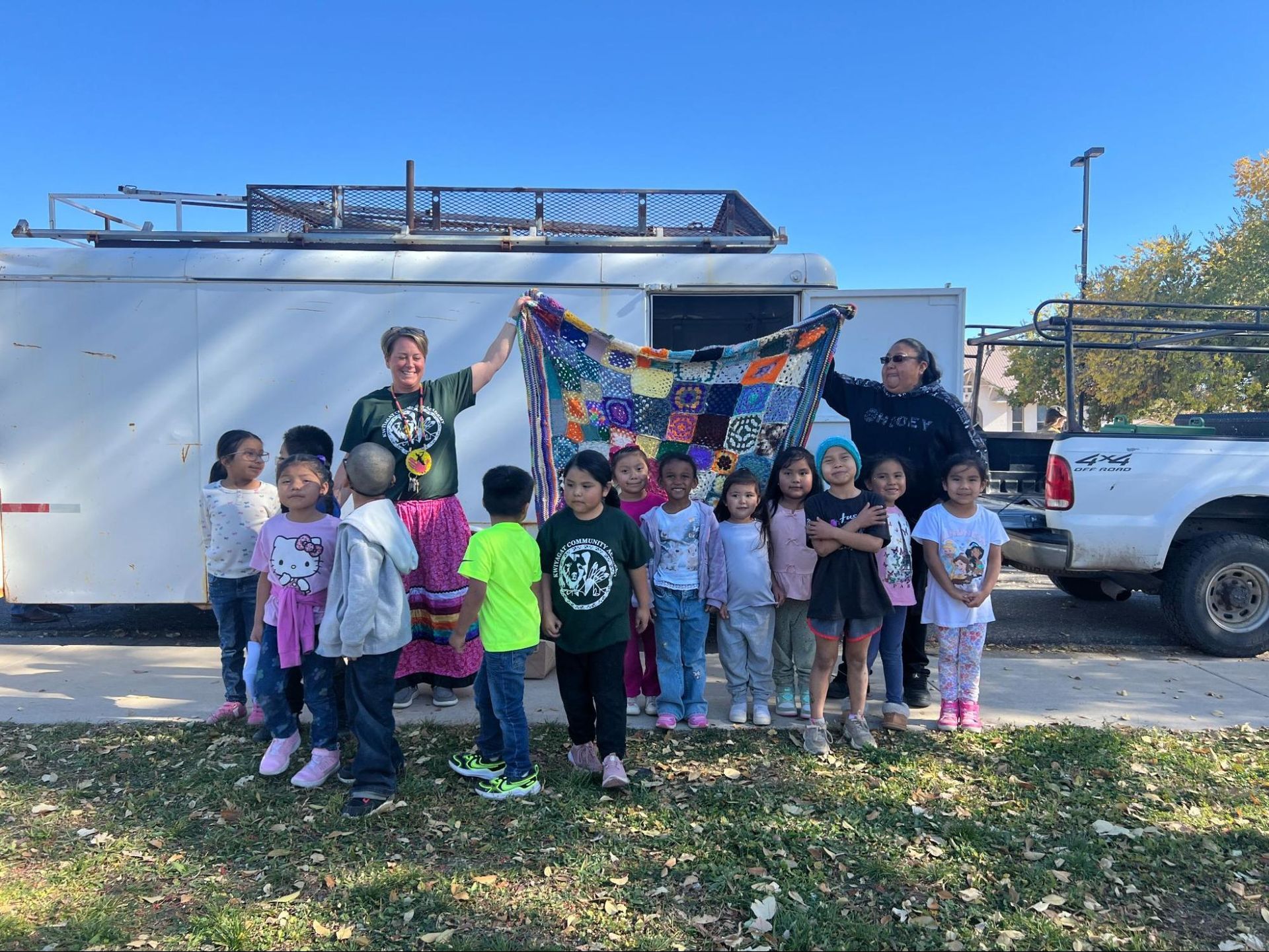 Group of children holding a colorful quilt outdoors beside a white truck and trailer