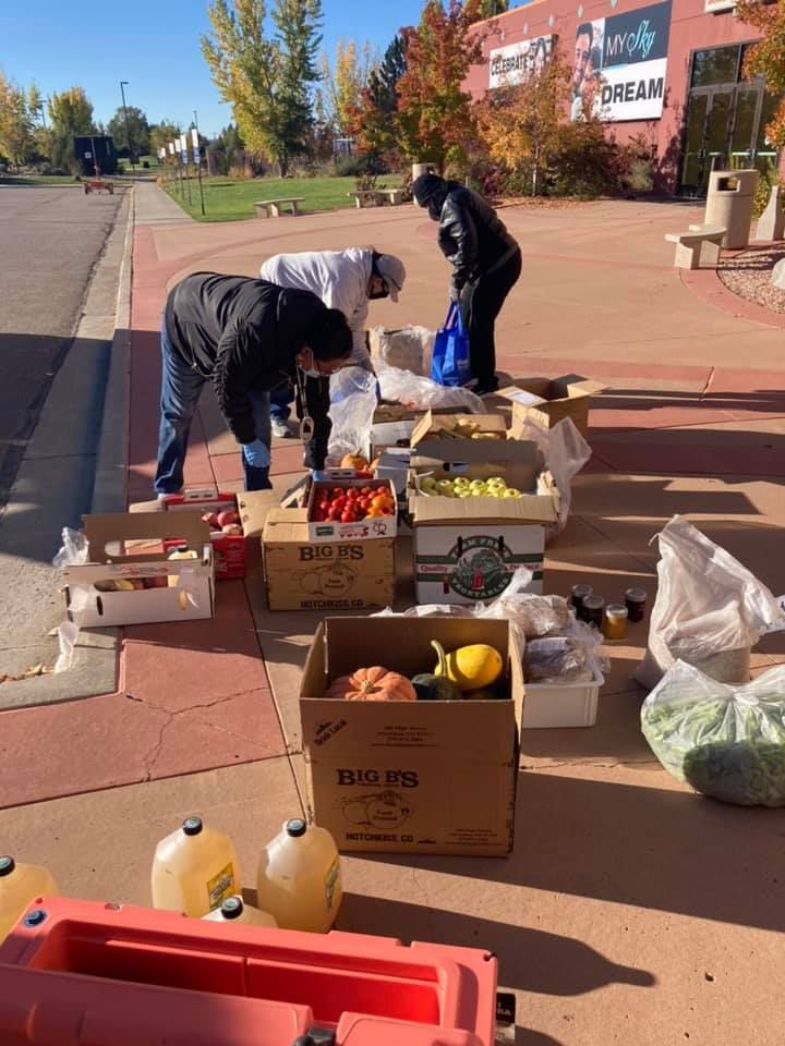 People sorting produce and goods on tables along a sunny sidewalk with boxes and jugs