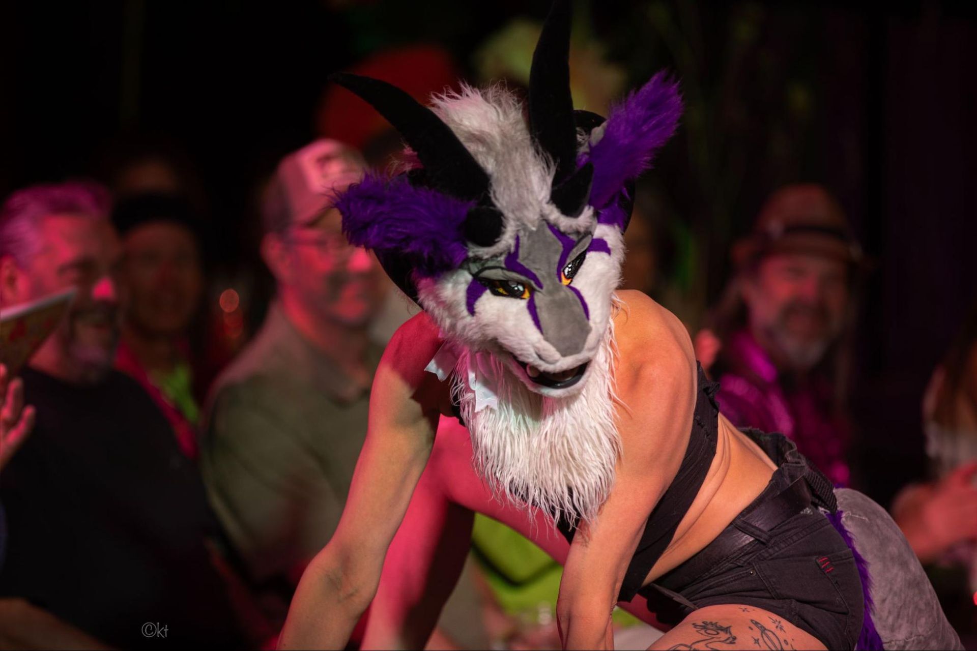 Masked performer in purple and black costume posing on stage under pink lights