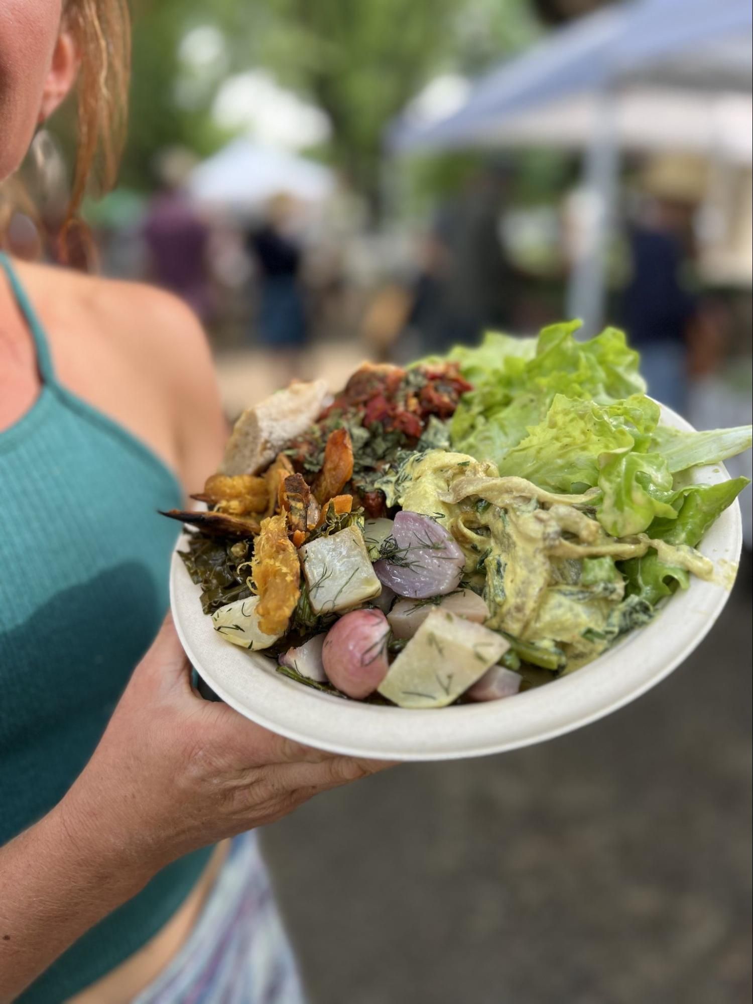 Person holding a plate of salad with greens, roasted vegetables, and a creamy dressing outdoors