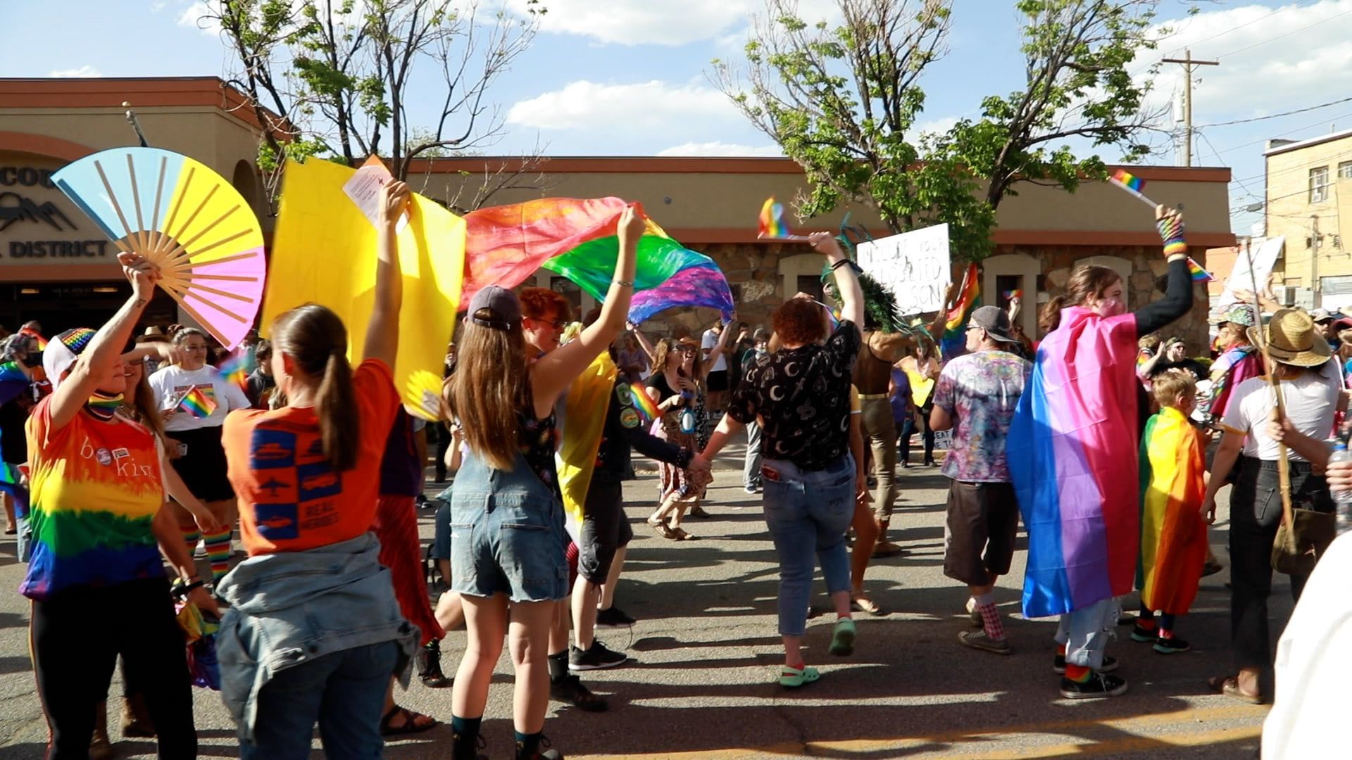 People waving colorful flags at an outdoor parade in front of buildings