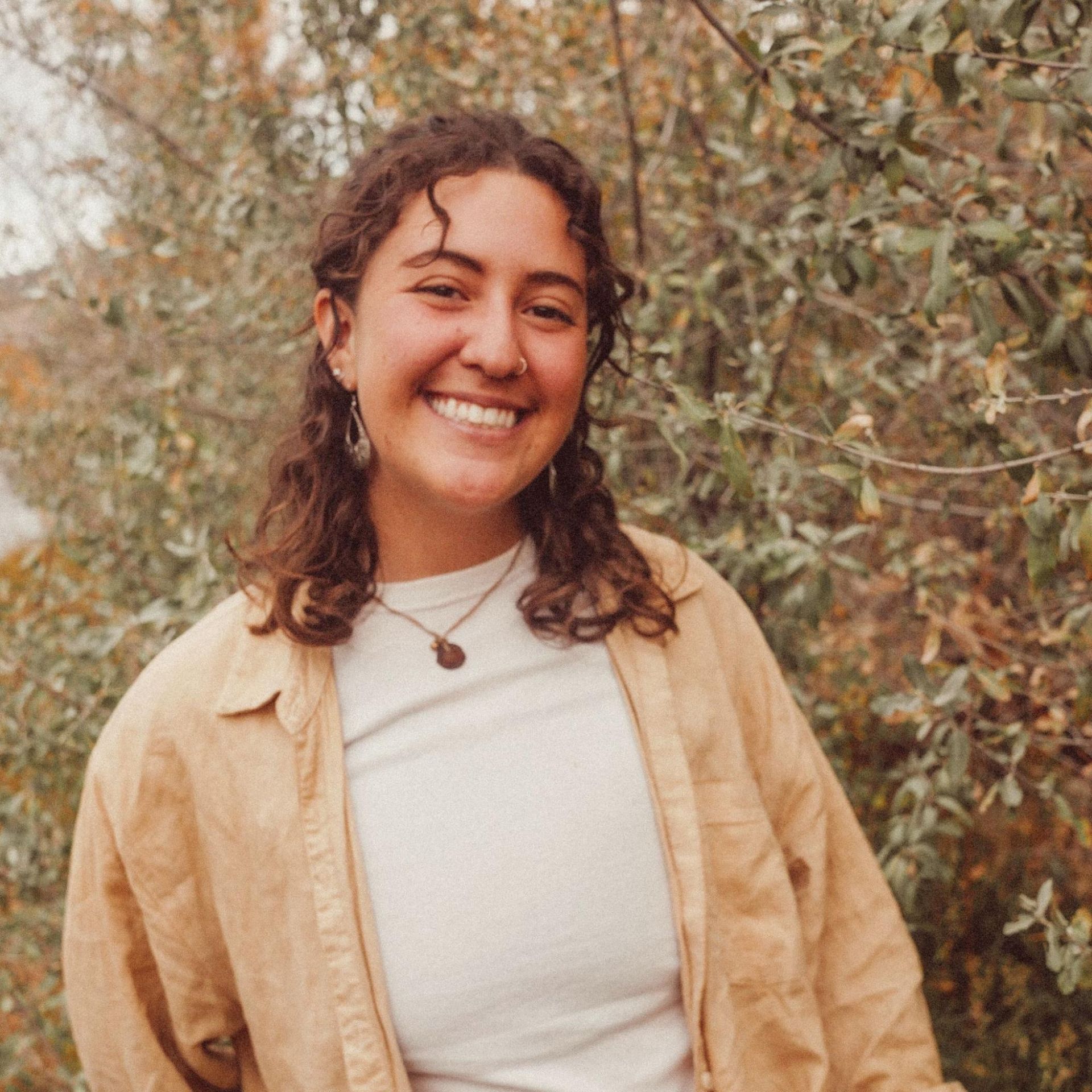 Smiling person in a beige shirt standing outdoors among trees and foliage